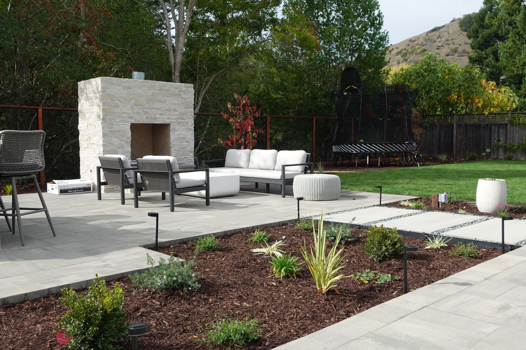 Paver patio with a stone fireplace, seating area, planting beds, lawn, and landscape lighting